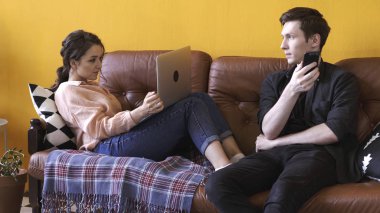 Dizüstü bilgisayar ve akıllı telefon kullanırken evde kahverengi deri kanepede birlikte oturan genç adam ve kadın yakın çekim. Stok görüntüleri. Evde çalışan bir çift serbest çalışan.