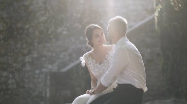 Gün ışığında oturan güzel yeni evliler. Eylem. Güzel gelin ve damat oturup birbirlerine parlak güneş ışığı ile aydınlatılmış bakmak. Gençlerin samimi ve şefkatli sevgisi