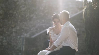 Gün ışığında oturan güzel yeni evliler. Eylem. Güzel gelin ve damat oturup birbirlerine parlak güneş ışığı ile aydınlatılmış bakmak. Gençlerin samimi ve şefkatli sevgisi