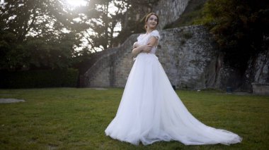 Ortaçağ duvarlarının arka planda güzel gelin. Eylem. Ağaçların arka planda yeşil çim üzerinde poz beyaz elbiseli Gelin. Gelin yeşil bahçede gelinlik şefkatle gülümsüyor
