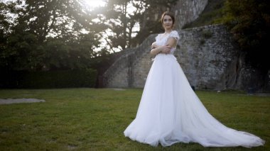 Ortaçağ duvarlarının arka planda güzel gelin. Eylem. Ağaçların arka planda yeşil çim üzerinde poz beyaz elbiseli Gelin. Gelin yeşil bahçede gelinlik şefkatle gülümsüyor