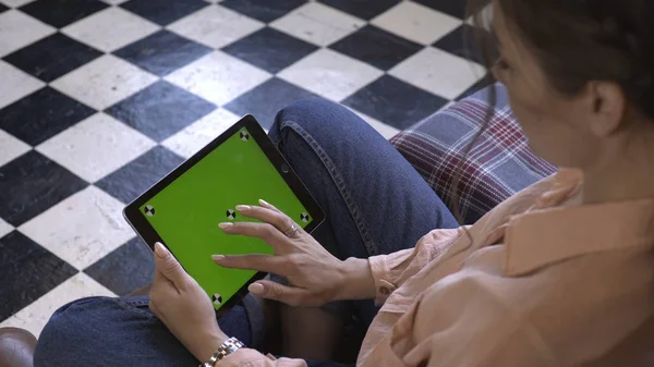 Vista de cerca de la hermosa mujer joven sentada en la silla y tocando en la tableta con la pantalla verde croma clave. Imágenes de archivo. Pantalla de clave de croma para la colocación de su propio contenido . — Foto de Stock