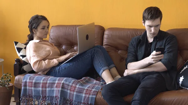 Primer plano de hombre y mujer jóvenes sentados juntos en el sofá de cuero marrón en casa mientras se utiliza el ordenador portátil y el teléfono inteligente. Imágenes de archivo. Pareja de freelancers trabajando en casa . — Foto de Stock