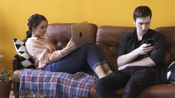 Nahaufnahme eines jungen Mannes und einer jungen Frau, die zusammen auf einer braunen Ledercouch zu Hause sitzen, während sie Laptop und Smartphone benutzen. Archivmaterial. Freiberufler, die zu Hause arbeiten. — Stockfoto