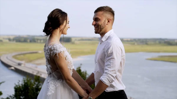 Newlyweds on background of fields and horizon with sky. Action. Husband and wife hold hands, look each other in eye and smile at background of landscape fields — Stock Photo, Image