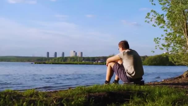 Młody człowiek siedzi na brzegu rzeki na tle miasta. Materiały stockowe. Człowiek z widokiem na naturę siedzi nad rzeką. Przyroda, fale na rzece i na tle można zobaczyć wysoki wzrost miasta — Wideo stockowe