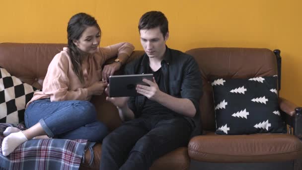 Vista de cerca de la feliz pareja joven sentada en el sofá en casa y mirando a la pantalla de la tableta. Imágenes de archivo. Hombre y mujer relajándose en el sofá, feliz fin de semana — Vídeo de stock