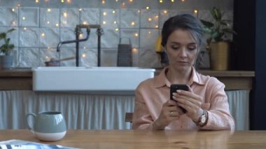 Ahşap masada mutfakta oturan ve akıllı telefon kullanarak pembe gömlek kahverengi saçlı güzel genç kadın yakından görünümü. Stok görüntüleri. Arka planda modern dekore edilmiş ev iç