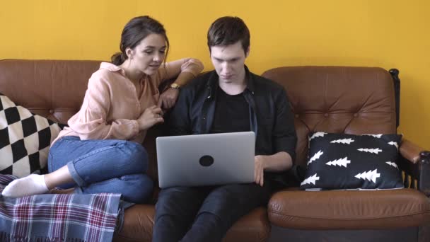Maravillosa pareja joven sentada en el sofá en casa y mirando a la pantalla del ordenador portátil. Imágenes de archivo. Hombre y mujer relajándose en el sofá y discutiendo su trabajo — Vídeo de stock