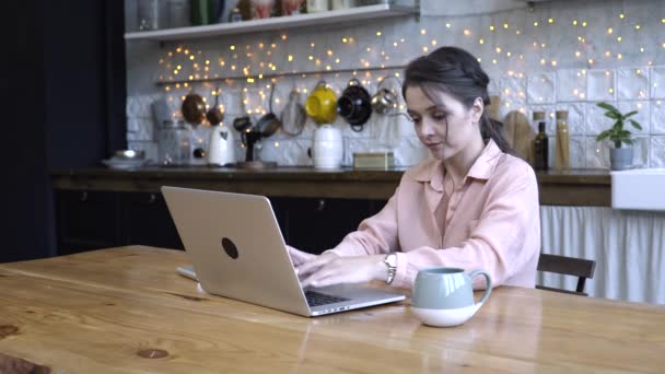 Konzentriert sitzt eine junge Frau in der Küche am Holztisch und tippt mit der Tasse Tee oder Kaffee auf ihrem persönlichen silbernen Laptop. Archivmaterial. modern dekorierte Innenausstattung auf dem Hintergrund — Stockvideo