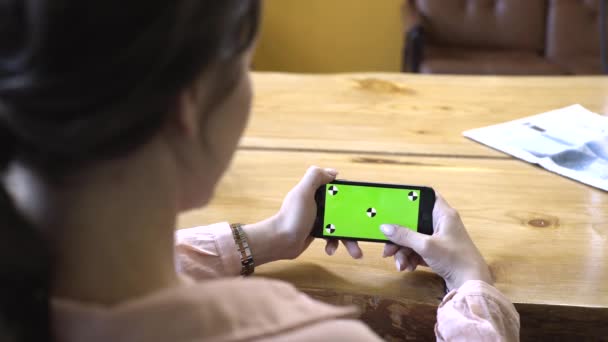 Vista da parte de trás da jovem mulher com cabelo castanho em camisa rosa sentado na mesa de madeira e segurando iPhone na mão batendo na tela verde chave chroma. Imagens de stock. Conceito de tecnologia — Vídeo de Stock