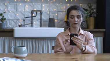 Ahşap masada mutfakta oturan ve akıllı telefon kullanarak pembe gömlek kahverengi saçlı güzel genç kadın yakından görünümü. Stok görüntüleri. Arka planda modern dekore edilmiş ev iç