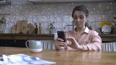 Pembe gömlek kahverengi saçlı güzel genç kadın ahşap masada mutfakta oturan ve akıllı telefon kullanarak. Stok görüntüleri. Arka planda modern dekore edilmiş ev iç