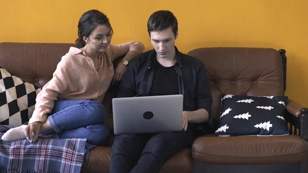 Magnifique jeune couple assis sur le canapé à la maison et regardant à l'écran d'ordinateur portable. Images d'archives. Homme et femme se relaxent sur le canapé et discutent de leur travail — Photo