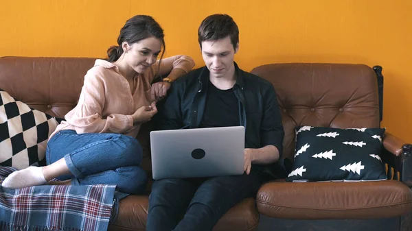 Maravillosa pareja joven sentada en el sofá en casa y mirando a la pantalla del ordenador portátil. Imágenes de archivo. Hombre y mujer relajándose en el sofá y discutiendo su trabajo — Foto de Stock