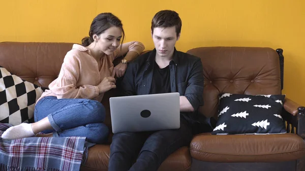 Maravillosa pareja joven sentada en el sofá en casa y mirando a la pantalla del ordenador portátil. Imágenes de archivo. Hombre y mujer relajándose en el sofá y discutiendo su trabajo — Foto de Stock