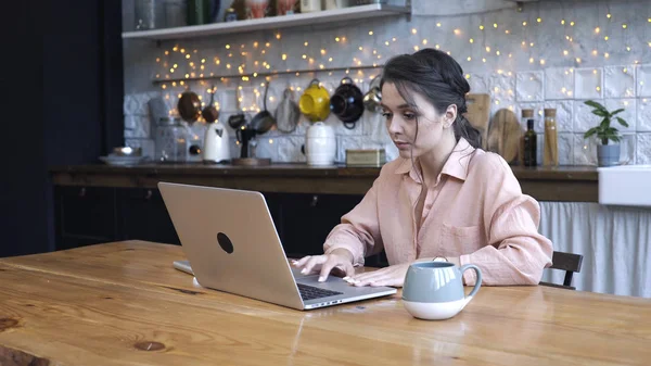Сконцентрированная молодая женщина, сидящая на кухне за деревянным столом и печатающая на своем личном серебряном ноутбуке с чашкой чая или кофе. Запись. Современный декорированный интерьер дома на заднем плане — стоковое фото