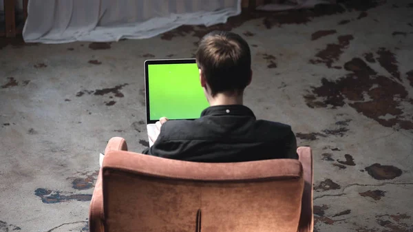 Widok z tyłu młodego mężczyzny w czarnej koszulce siedzącej w brązowym wygodnym fotelu i pracy na laptopie z kluczem chrominancji zielony ekran. Materiały stockowe. Miejsce własnej treści. — Zdjęcie stockowe