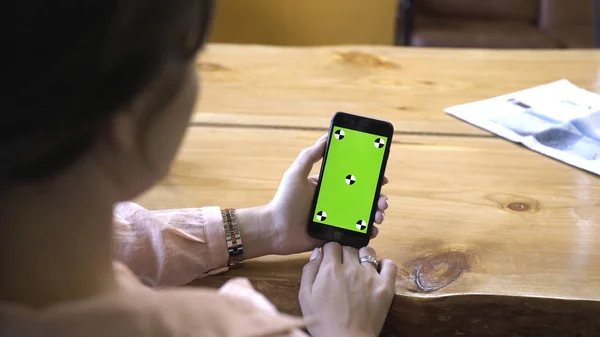 Mujer joven con el pelo castaño en camisa rosa sentada en la mesa de madera, sosteniendo el iPhone en la mano y mirando a la pantalla verde croma clave. Imágenes de archivo. Concepto de nueva tecnología Smartphone — Foto de Stock