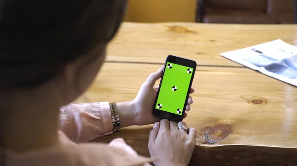 View from the shoulder of woman with brown hair in pink shirt holding iPhone in hand and looking at the chroma key green screen. Stock footage. Cellphone with chroma key screen