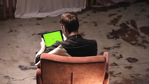 Vista da parte de trás do jovem com cabelo castanho segurando computador tablet preto com tela verde enquanto sentado no sofá em casa. Imagens de stock. Chave Chroma — Fotografia de Stock