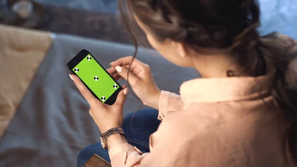 Mulher bonita com cabelo castanho sentado em casa em camisa rosa e deslizando em seu smartphone na tela verde chave chroma. Imagens de stock. Smartphone novo conceito de tecnologia — Fotografia de Stock