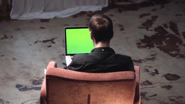 Blick von hinten auf einen jungen Mann in schwarzem Hemd, der im braunen bequemen Stuhl sitzt und an einem Laptop mit grünem Bildschirm arbeitet. Archivmaterial. Ort der eigenen Inhalte. — Stockvideo