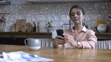 Pembe gömlek kahverengi saçlı güzel genç kadın ahşap masada mutfakta oturan ve akıllı telefon kullanarak. Stok görüntüleri. Arka planda modern dekore edilmiş ev iç