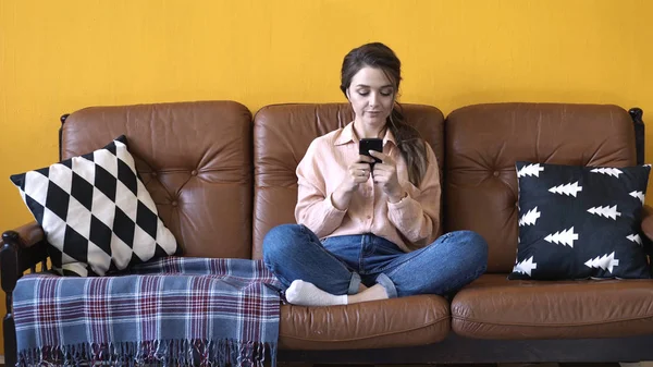 Vista da jovem mulher bonita em camisa rosa e jeans sentado no sofá marrom com travesseiros e digitando em seu smartphone. Imagens de stock. Mulher relaxante em casa — Fotografia de Stock