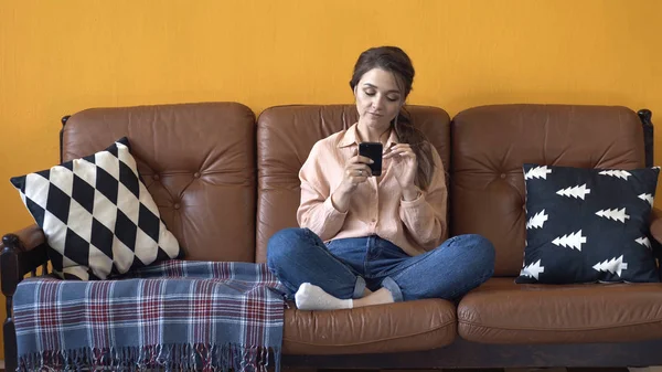 Happy young woman with dark hair in pink shirt and jeans sitting on the brown couch with pillows and typing on her smartphone. Stock footage. Woman relaxing at home — Stock Photo, Image