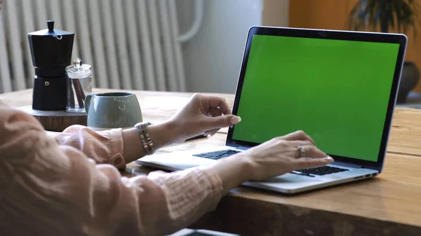 Mujer joven en camisa rosa sentada en la gran mesa de madera y escribiendo en su computadora portátil con pantalla verde croma clave. Imágenes de archivo. Pantalla de clave de croma para la colocación de su propio contenido . — Foto de Stock