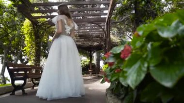 Genç güzel gelin bahçede beyaz elbise standı. Eylem. Gazebo altında yeşil bahçede duran narin beyaz elbiseli genç gelinin arka görünümü