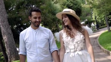 Yeni evliler gülümsüyor ve parkta el ele tutuşuyorlar. Eylem. Güzel genç çift balayı mutluluk ile ışınlanıyor. Şapkalı genç kadın kocasına gülümsüyor