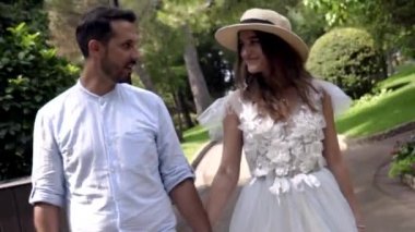 Yeni evliler gülümsüyor ve parkta el ele tutuşuyorlar. Eylem. Güzel genç çift balayı mutluluk ile ışınlanıyor. Şapkalı genç kadın kocasına gülümsüyor