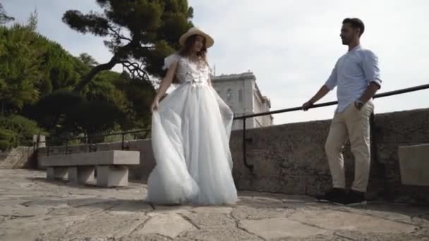 Belo jovem casal em lua de mel. Acção. Marido admira sua bela jovem esposa girando em vestido branco. Recém-casados no fundo do deck de observação de pedra — Vídeo de Stock