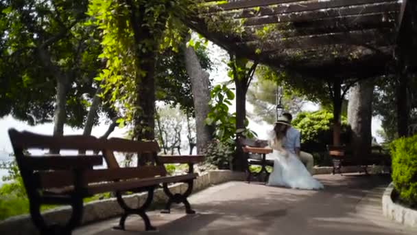 美しいガゼボのベンチに座っているカップル。アクション。ハネムーンでガゼボに座っている美しいカップル。夫は大切な妻の頬にキスをする。甘くてロマンチックなカップル — ストック動画