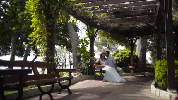 Casal sentado no banco em belo mirante. Acção. Belo casal sentado no gazebo em lua de mel. Marido beija sua preciosa esposa na bochecha. Doce e romântico casal — Vídeo de Stock