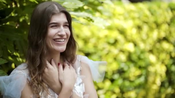 Mujer hermosa joven posando en vestido blanco sobre fondo de arbustos verdes. Acción. Hermosa apariencia delicada de mujer joven. Sesión de fotos suave de verano de la mujer joven — Vídeo de stock