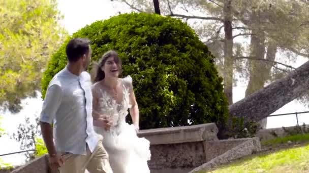Newlyweds run in park in summer. Action. Beautiful loving couple romantically run away holding hands in park. Wife in white dress holding her husbands hand illuminated by sunlight — Stock Video