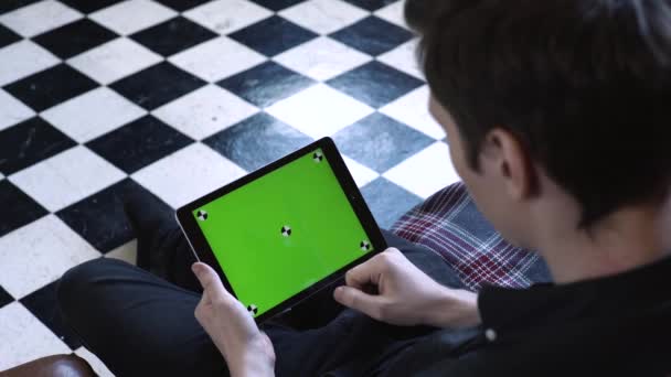 Visão superior do jovem em camisa preta sentado no sofá em casa e tocando na tela verde do tablet chroma key. Imagens de stock. Novo conceito de tecnologia — Vídeo de Stock