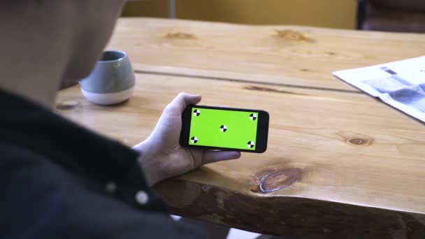 View from the shoulder of young man in black shirt sitting at wooden table and looking at phone chroma key green screen at home. Stock footage. Smartphone new technology concept — Stock Video