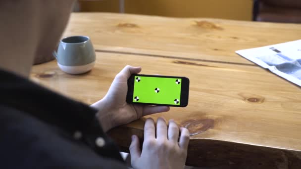 나무 테이블에 앉아 집에서 전화 크로마 키 녹색 화면을보고 검은 셔츠에 젊은 남자의 어깨에서보기. 스톡 푸티지. 스마트폰 신기술 컨셉 — 비디오