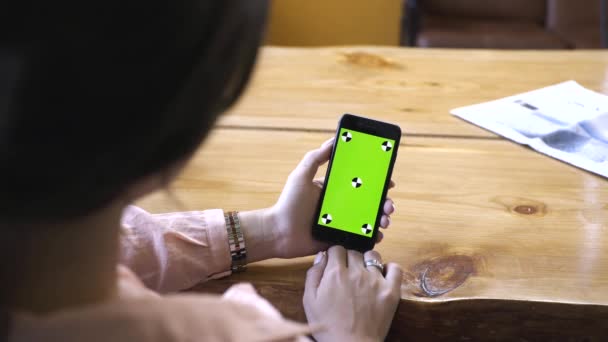 Mulher jovem com cabelos castanhos em camisa rosa sentado na mesa de madeira, segurando iPhone na mão e olhando para a tela verde chave chroma. Imagens de stock. Smartphone novo conceito de tecnologia — Vídeo de Stock