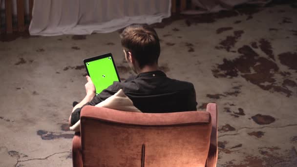 Θέα από το πίσω μέρος του νεαρού άνδρα με καστανά μαλλιά κρατώντας μαύρο tablet υπολογιστή με πράσινη οθόνη, ενώ κάθεται στον καναπέ στο σπίτι. Υλικό από μετοχές. Πλήκτρο αποχρώσεων — Αρχείο Βίντεο
