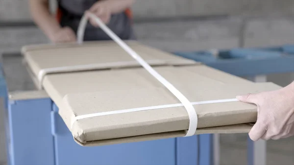 Primer plano de los trabajadores empaquetado paquete plano. Acción. Los trabajadores en overoles empaquetan material de construcción de fábrica para la entrega a domicilio. Los materiales de construcción o los muebles se entregan directamente de fábrica —  Fotos de Stock