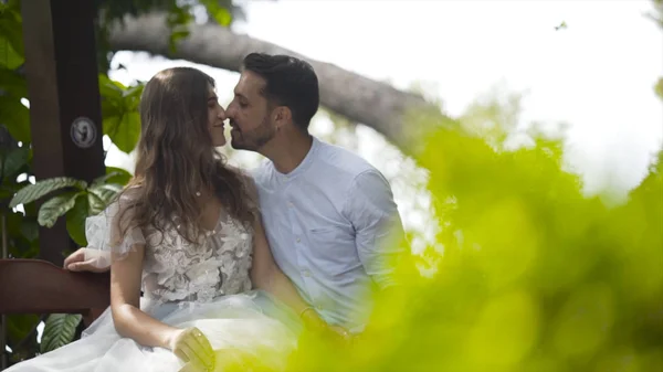 Junges schönes Paar küsst sich auf Bank im Garten. Aktion. junges Ehepaar küsst sich sanft auf Bank im grünen Garten. Frischvermählte entspannen im Garten — Stockfoto