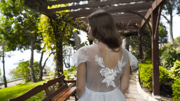 Young beautiful bride stand in white dress in garden. Action. Rear view of young bride in delicate white dress standing in green garden under gazebo.