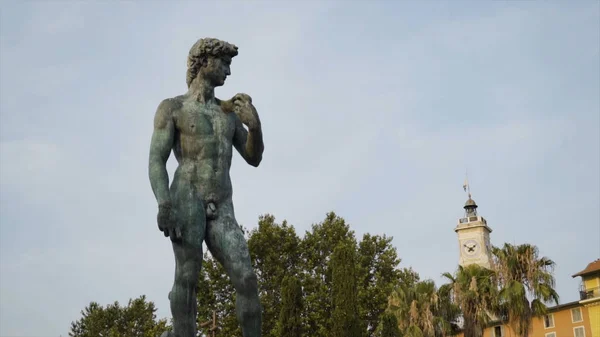 Statue of David in park. Action. Beautiful manly statue of David stands in park on background of sky. Nude statue of David is made of dark stone and stands on pedestal in park