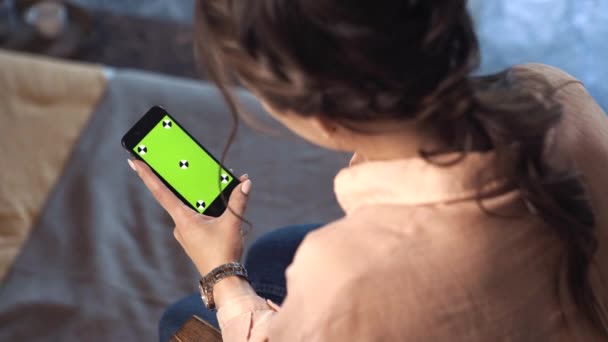 Hübsche Frau mit braunen Haaren sitzt zu Hause im rosafarbenen Hemd und schiebt auf ihrem Smartphone am grünen Bildschirm der Chroma-Taste. Archivmaterial. Smartphone neues Technologie-Konzept — Stockvideo