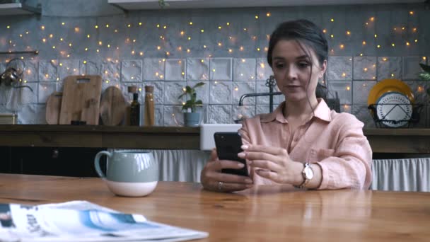 木製のテーブルで台所に座って、スマートフォンを使用してピンクのシャツで茶色の髪を持つかなり若い女性のクローズアップビュー。ストック映像。背景にモダンな内装の家のインテリア — ストック動画